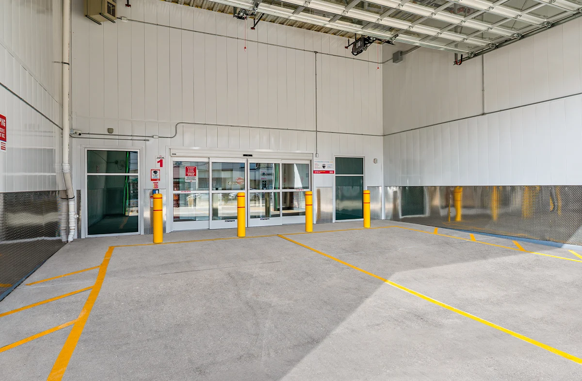 An interior view of a modern self-storage facility hallway with an open unit visible on the left, featuring a bright red roll-up door, corrugated metal walls, and a blue ADA accessibility sign.