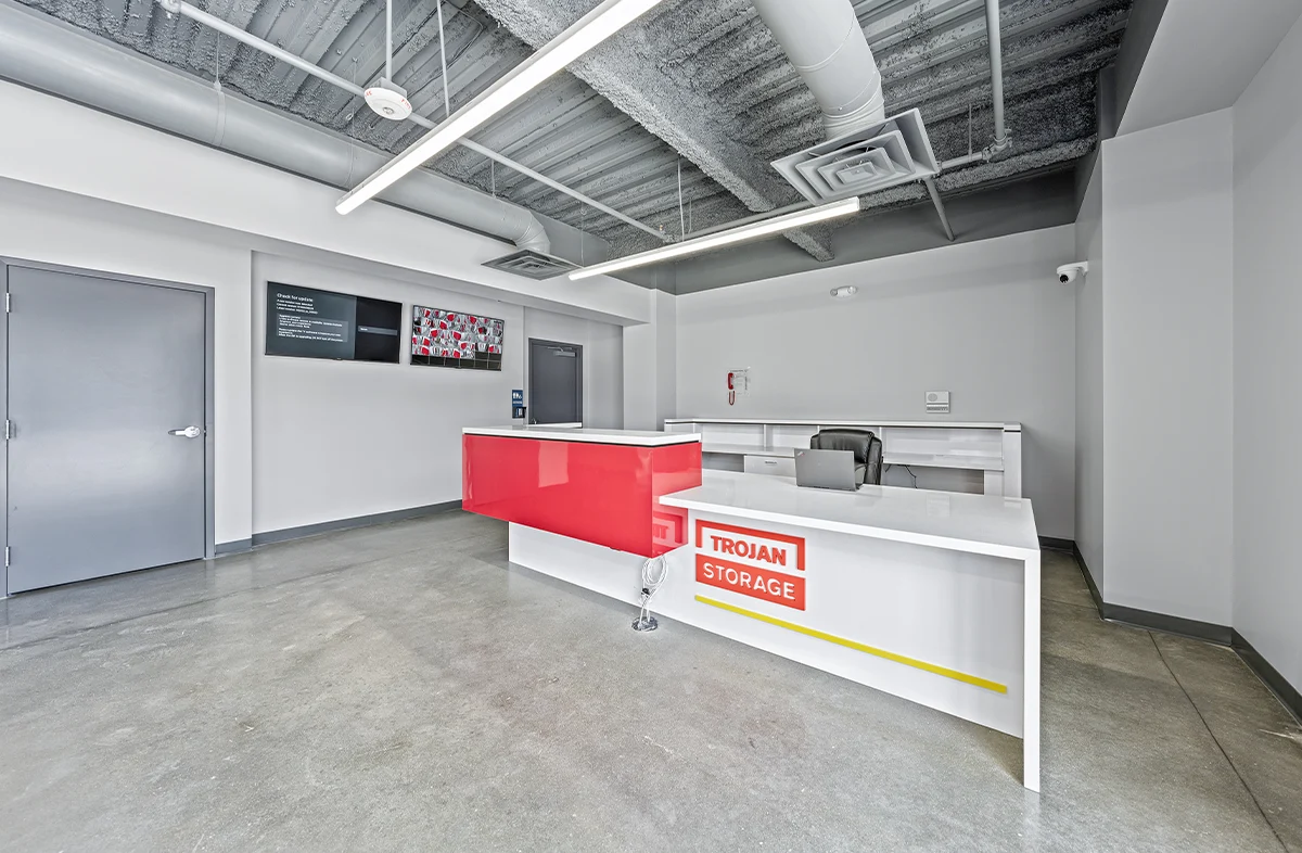 The modern, clean office lobby of a self-storage facility features a white and bright red reception desk with "Trojan Storage" branding, a concrete floor, and gray walls with monitors and a closed door.