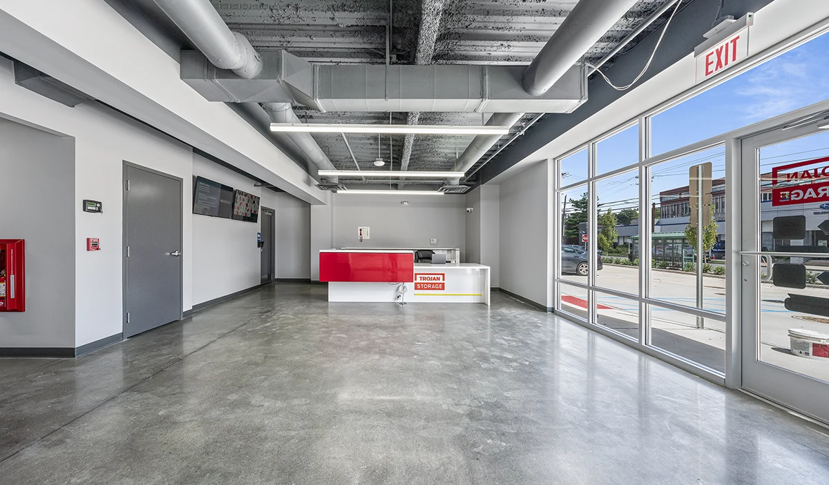 The modern, clean office lobby of a self-storage facility features a white and red reception desk, a concrete floor, gray walls, and bright lighting, with large windows overlooking the street and an "EXIT" sign above a glass door.
