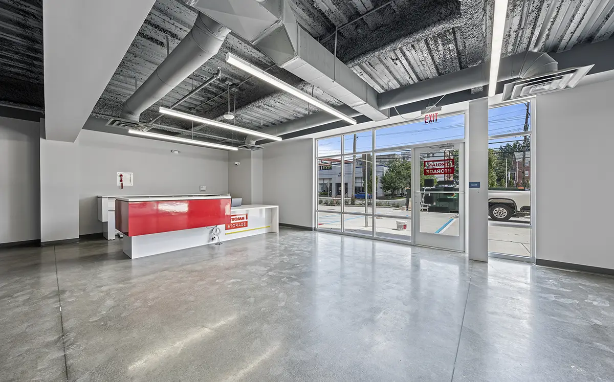 An interior hallway of a self-storage facility features a concrete floor, a bare metal ceiling with pipes and lights, and a white divider wall with an open storage unit that has a corrugated silver interior and a red roll-up door.