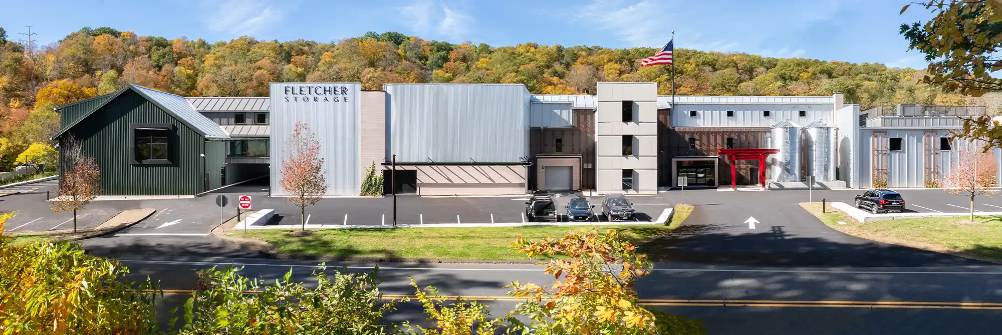 An aerial, panoramic shot of a modern, multi-building self-storage facility called "Fletcher Storage" set against a backdrop of colorful autumn trees. The facility has varied facades, including vertical metal siding and a small red torii-like gate entrance.