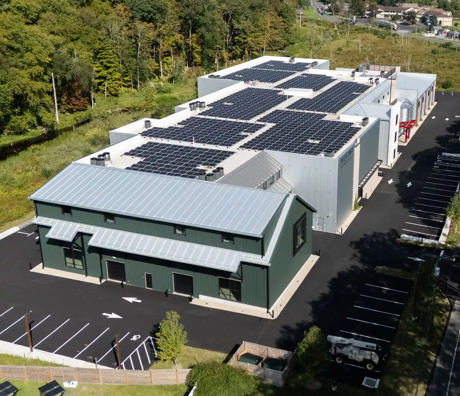 An aerial view of the Fletcher Storage facility's rooftop, which is almost completely covered in black solar panels. The building has a mixed metal and dark green facade and a paved parking lot surrounding it.