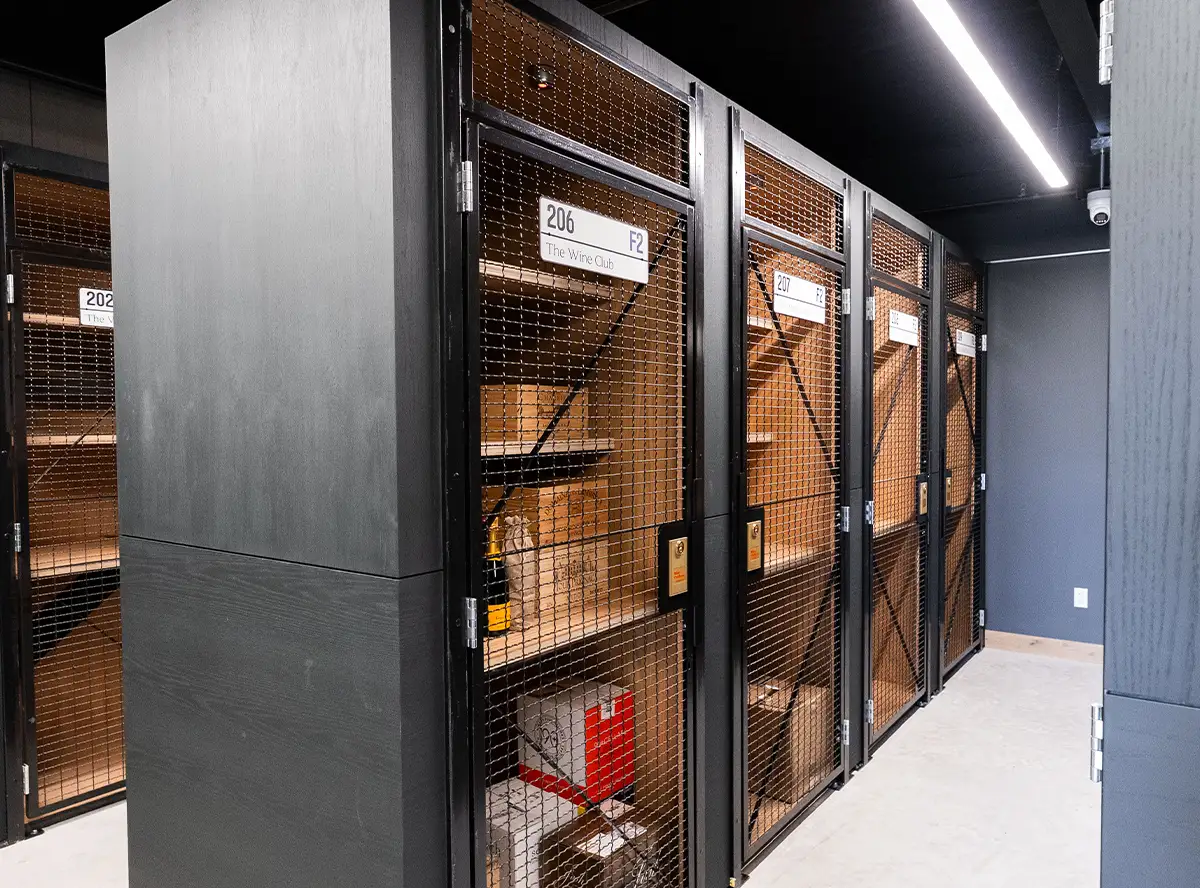 A row of climate-controlled wine storage units features dark gray wood paneling and black wire mesh doors with unit numbers like "206 The Wine Club," secured with visible yellow padlocks.