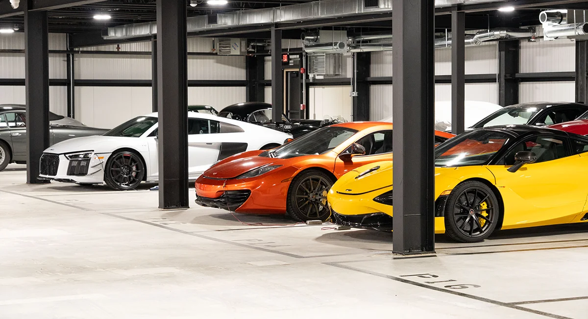 A luxury car storage area with a polished concrete floor and black support columns. Several high-end sports cars, including a white, orange, and yellow one, are parked in marked spaces.