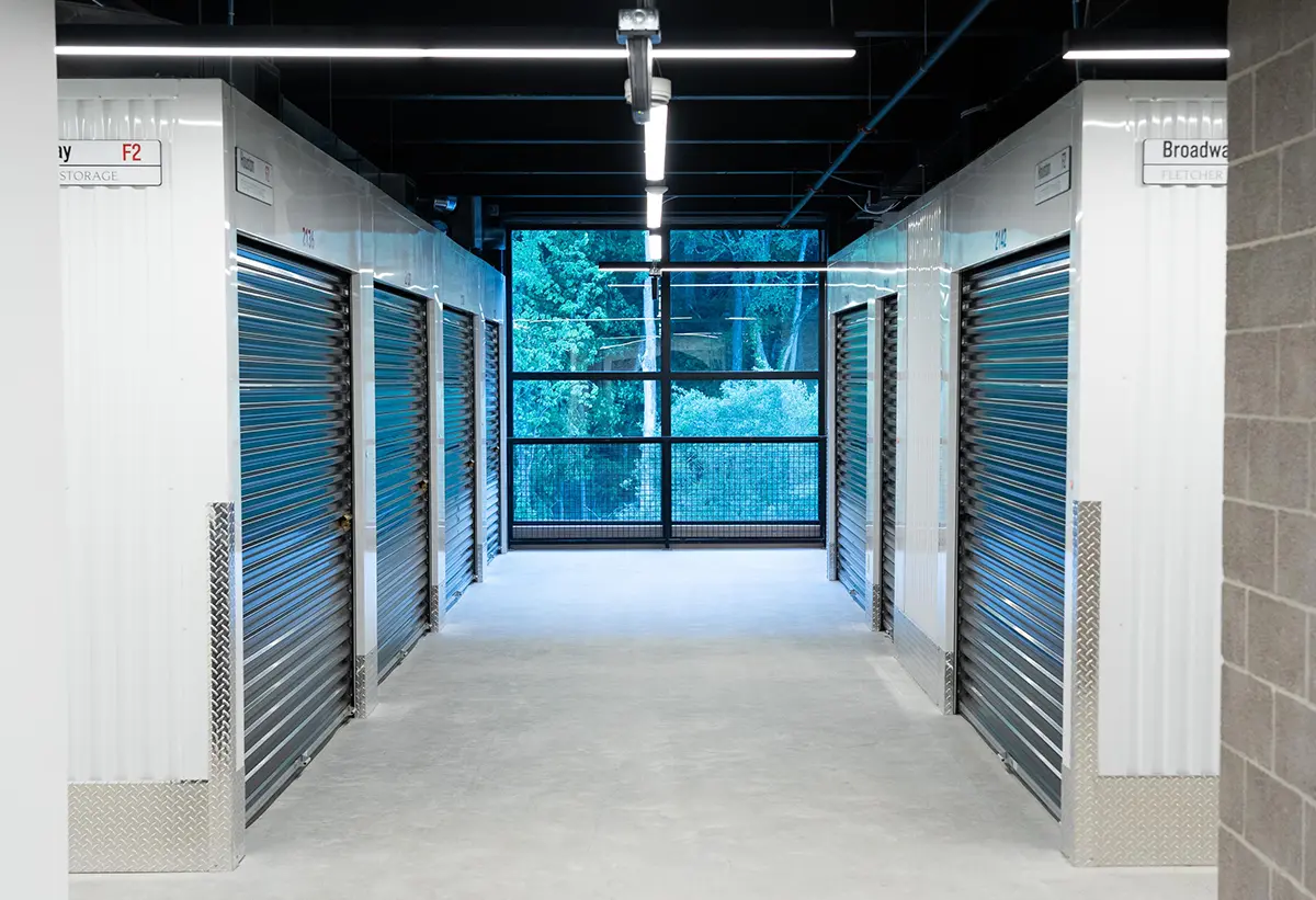 An interior view of a self-storage hallway with a concrete floor and a wide, open metal window in the distance, framed by storage units with horizontal blue-gray roll-up doors and white corrugated walls.