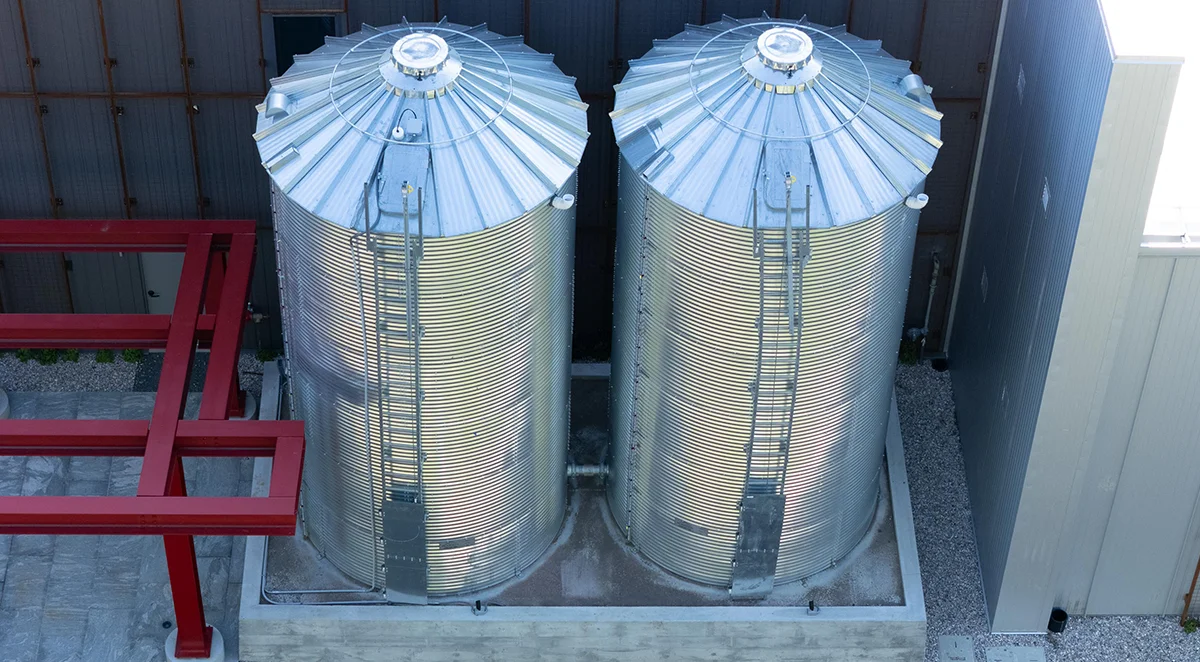 Two large, galvanized steel grain silos standing outdoors on a concrete base, indicating commercial storage or farm facilities.