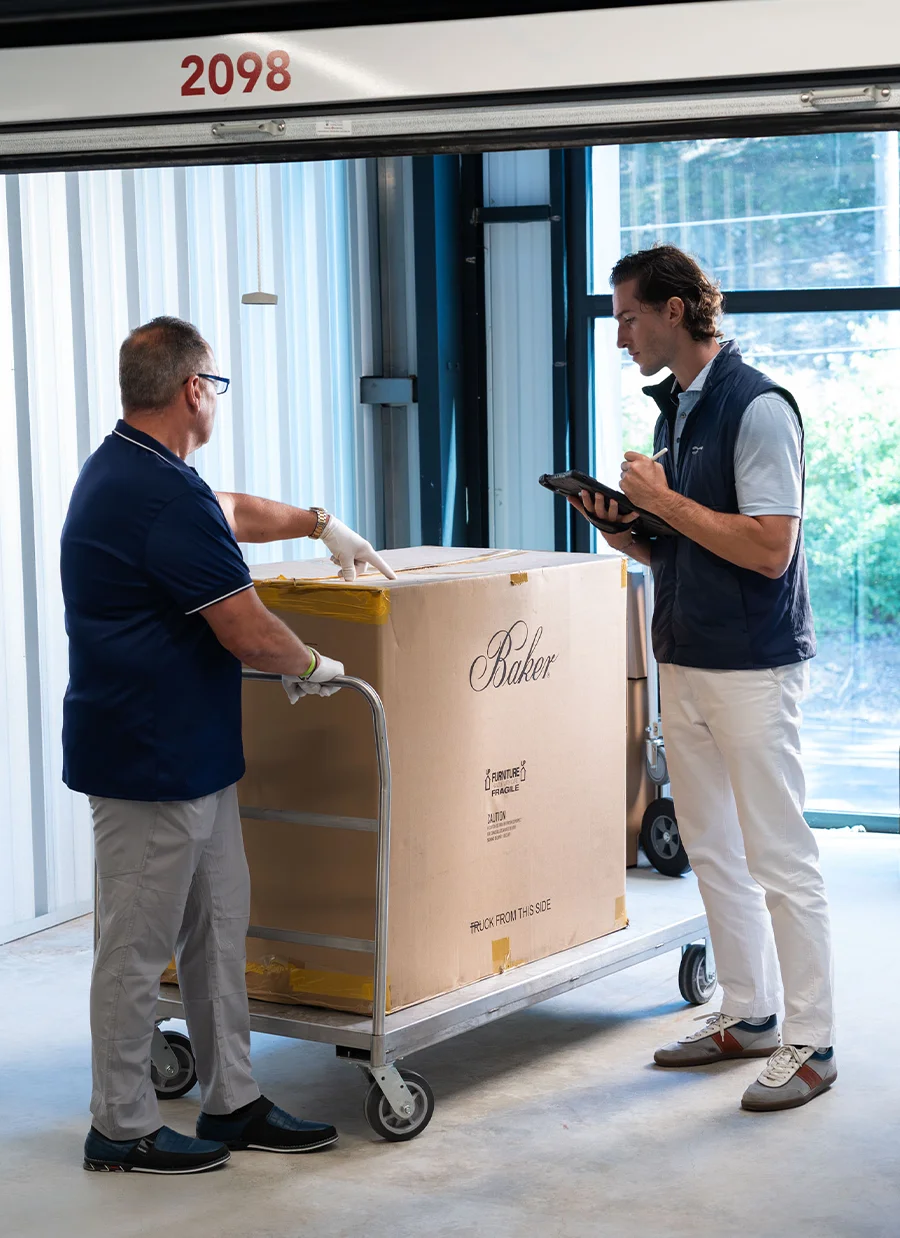 Two men handling a large, branded Baker Furniture moving box on a dolly, loading it into a self-storage unit.