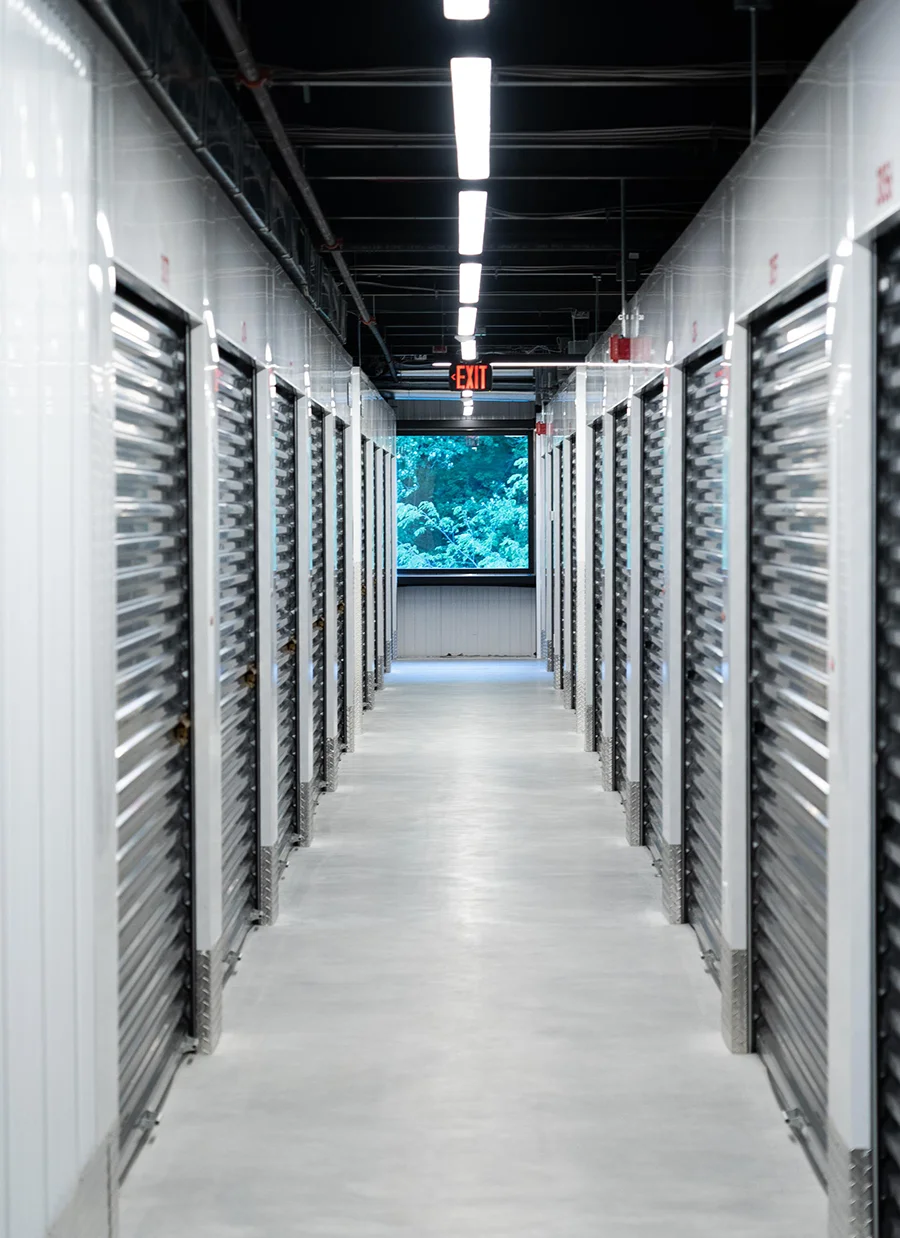 Long, brightly lit interior hallway of a self-storage facility with roll-up doors and a window view of green trees at the far end.
