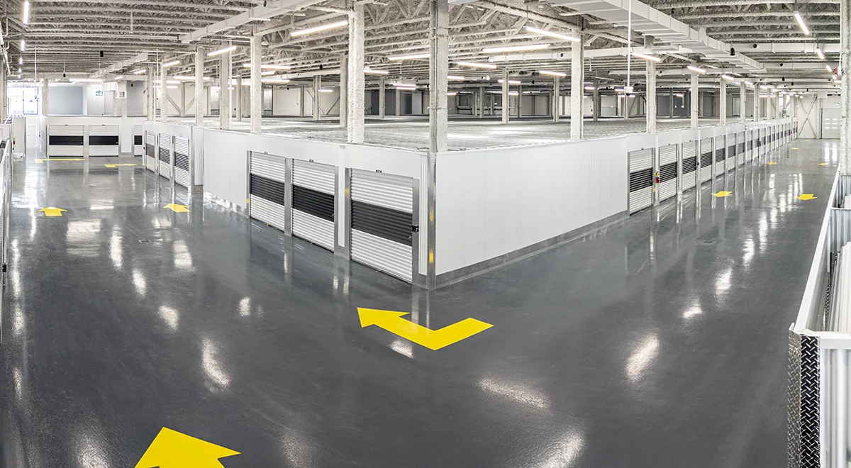 An overhead view of a large, brightly lit warehouse interior with a glossy gray floor and white walls, featuring rows of black and silver roll-up self-storage doors and yellow directional arrows painted on the floor.
