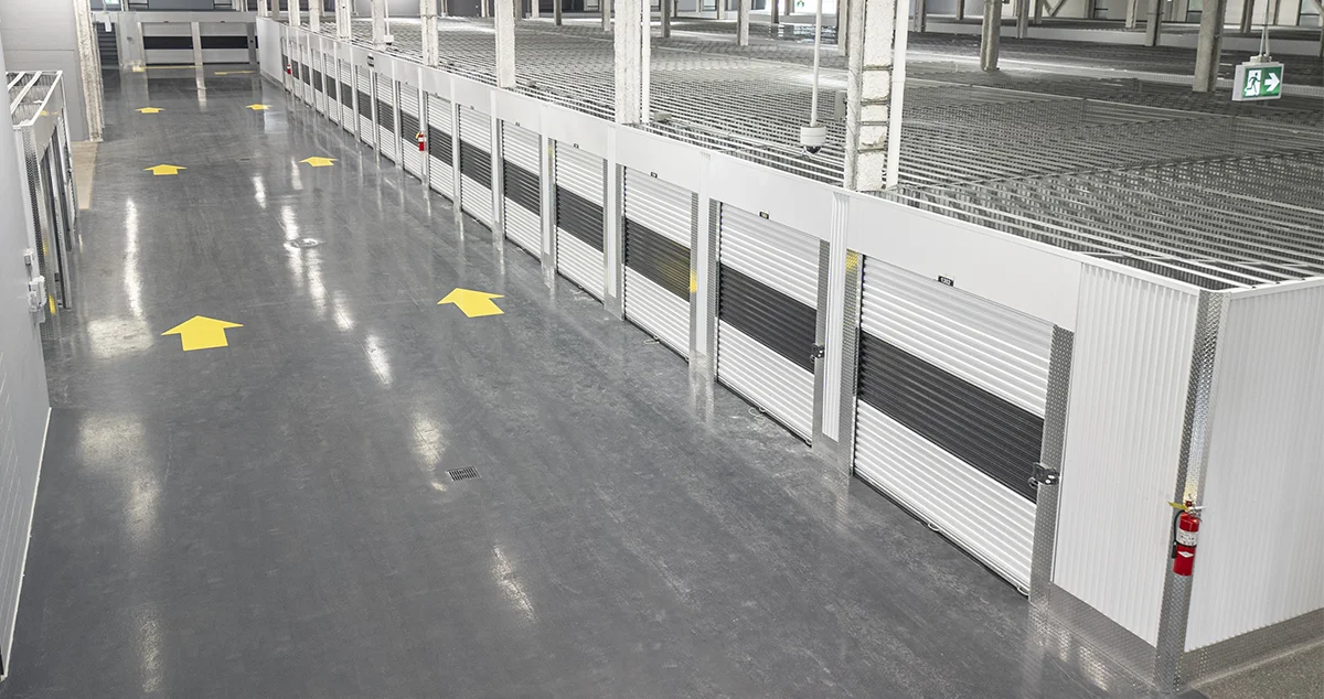 A wide-angle view of a vast, modern self-storage interior with a glossy gray floor, white walls, black and silver storage units, and yellow directional arrows painted on the floor.