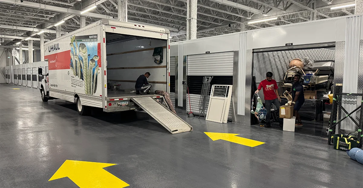 Inside a modern, climate-controlled self-storage facility, three men unload items from a U-Haul truck into an open storage unit, with yellow directional arrows painted on the gray floor.