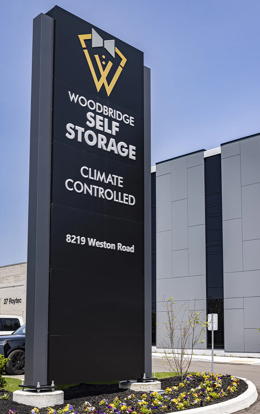 A prominent black vertical monument sign for "Woodbridge Self Storage, Climate Controlled" at 8219 Weston Road, featuring a yellow logo and set in front of the modern gray and black facility.