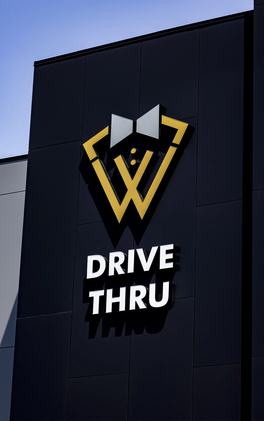 A close-up of the "W" logo for Woodbridge Self Storage, stylized as a bow tie, next to the white text "DRIVE THRU" on the black exterior of the building.