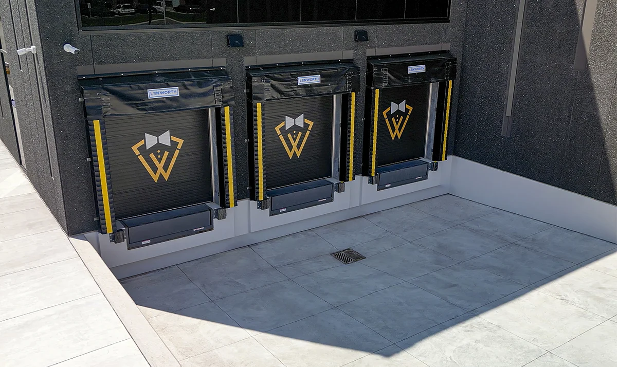 Three vertical, black roll-up dock doors, each featuring the gold and white "W" bow-tie logo of Woodbridge Self Storage, are set into the dark exterior wall of the facility.