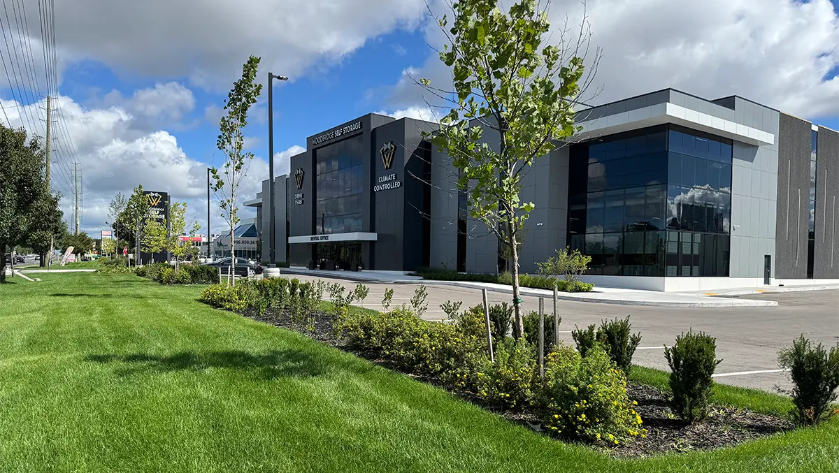 A wide outdoor view of the modern, gray and black Woodbridge Self Storage building with its yellow bow-tie logo, a prominent vertical sign, and a landscaped lawn and sidewalk in the foreground.