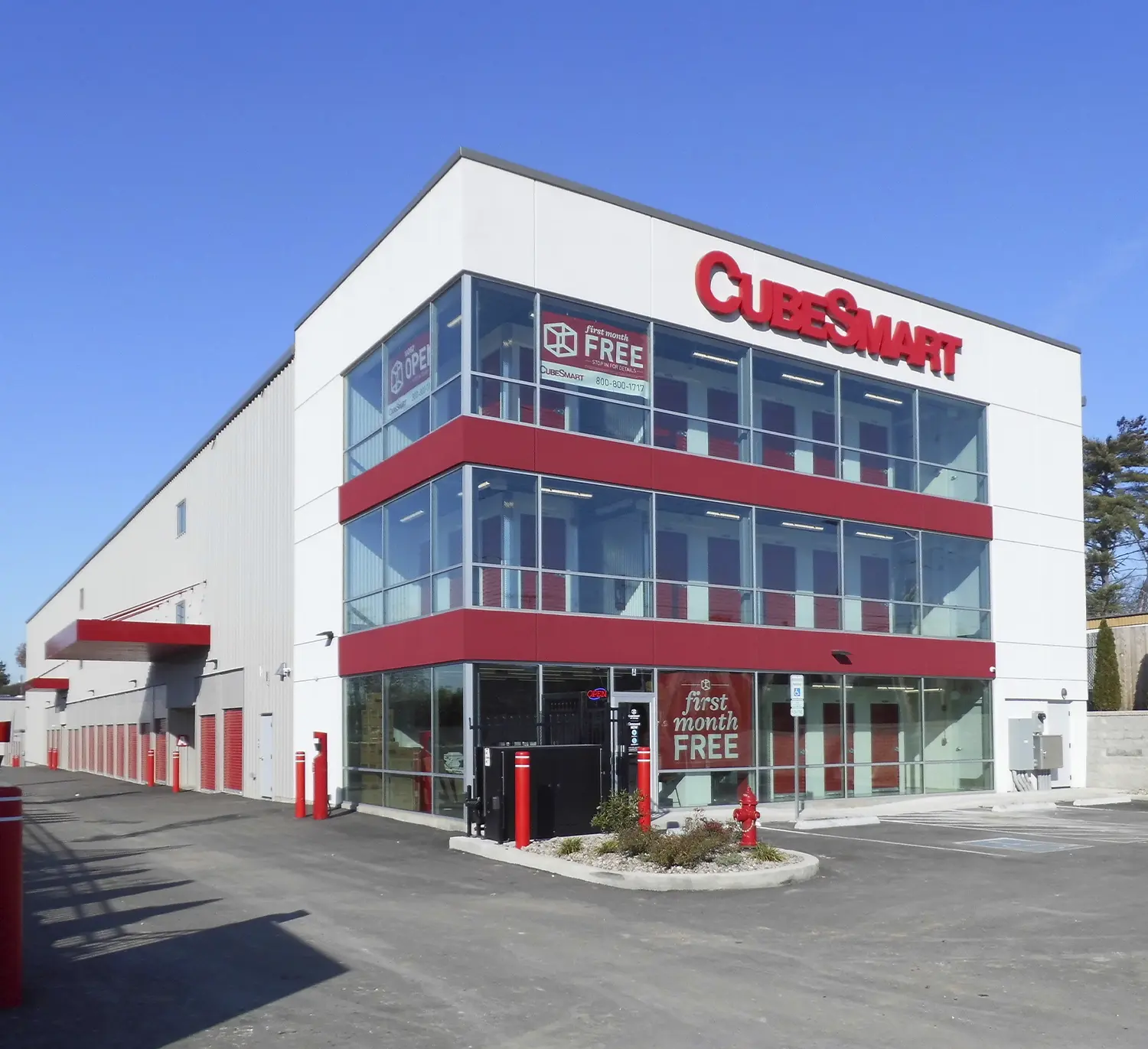 An exterior view of a modern CubeSmart self-storage facility with a white facade, red accents, and large windows. The building has an enclosed exterior drive-up area with red-doored units on the left.