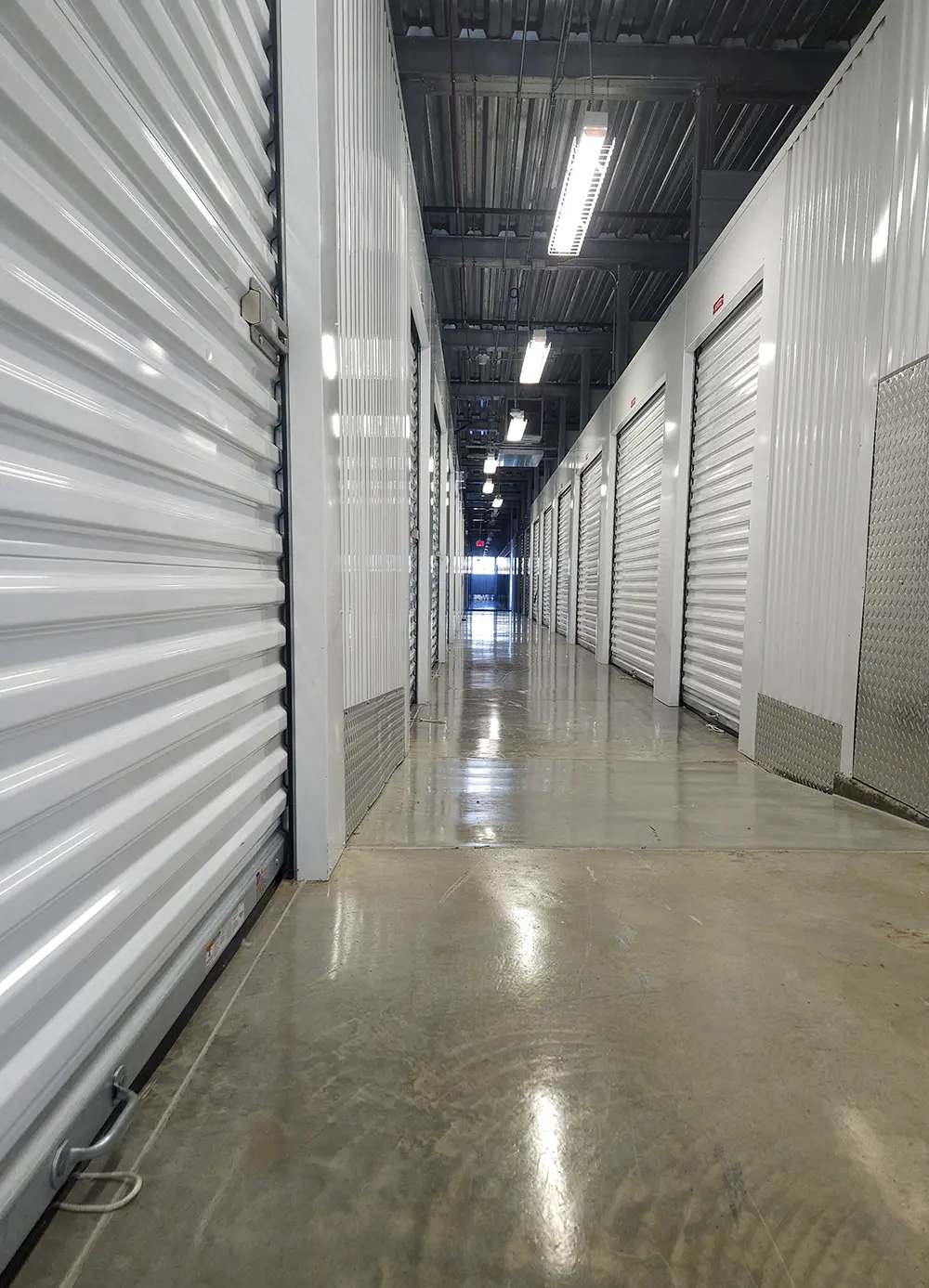 A brightly lit interior hallway of a self-storage facility with a concrete floor, corrugated white metal walls on both sides, and a series of white roll-up unit doors receding into the distance.