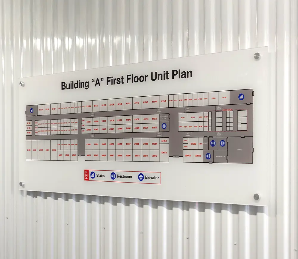 A clear acrylic sign titled "Building 'A' First Floor Unit Plan" is mounted on a white corrugated wall, displaying a detailed floor plan with a key for Stairs, Restroom, and Elevator.