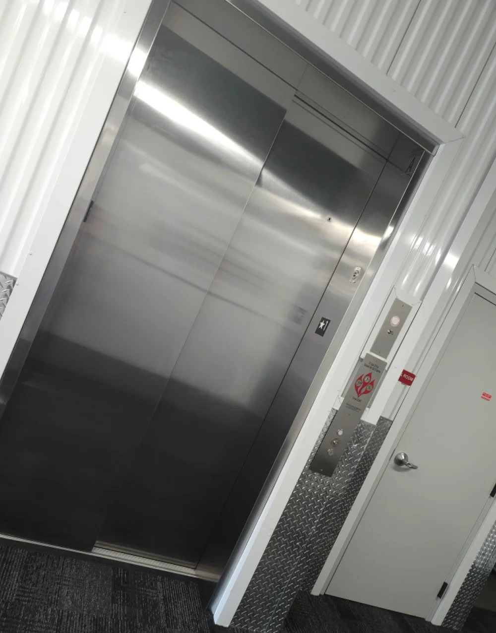 A stainless steel elevator door in a self-storage facility, with a floor selection panel, a key slot, and a red fire alarm sign mounted on the white corrugated wall next to it.