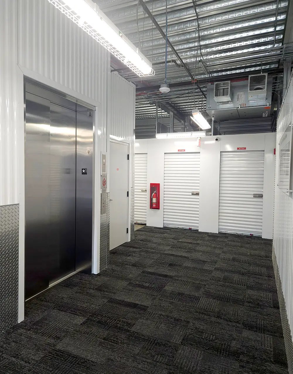 An interior view of a modern self-storage facility hallway with a patterned gray carpet floor. The space features white corrugated metal walls, bright overhead lighting, and a stainless steel elevator door next to a closed white door.