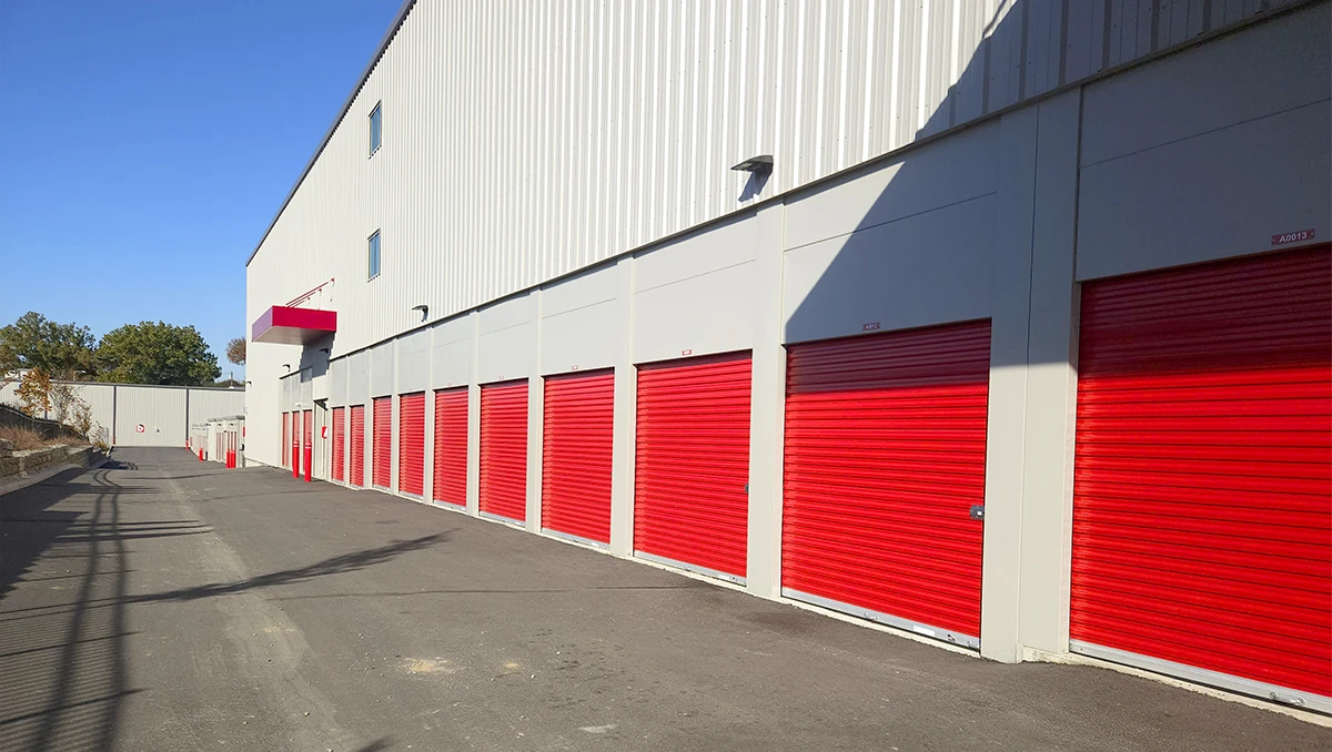 A ground-level, exterior view of a long row of outdoor self-storage units with bright red roll-up doors against a white corrugated metal building facade on a sunny day.