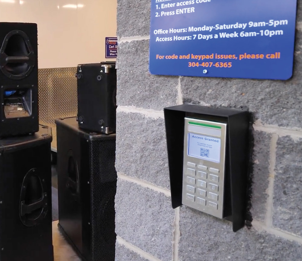 A digital keypad mounted on a cinder block wall displays "Access Granted" with a QR code. Large black speakers are stacked nearby in the storage area.