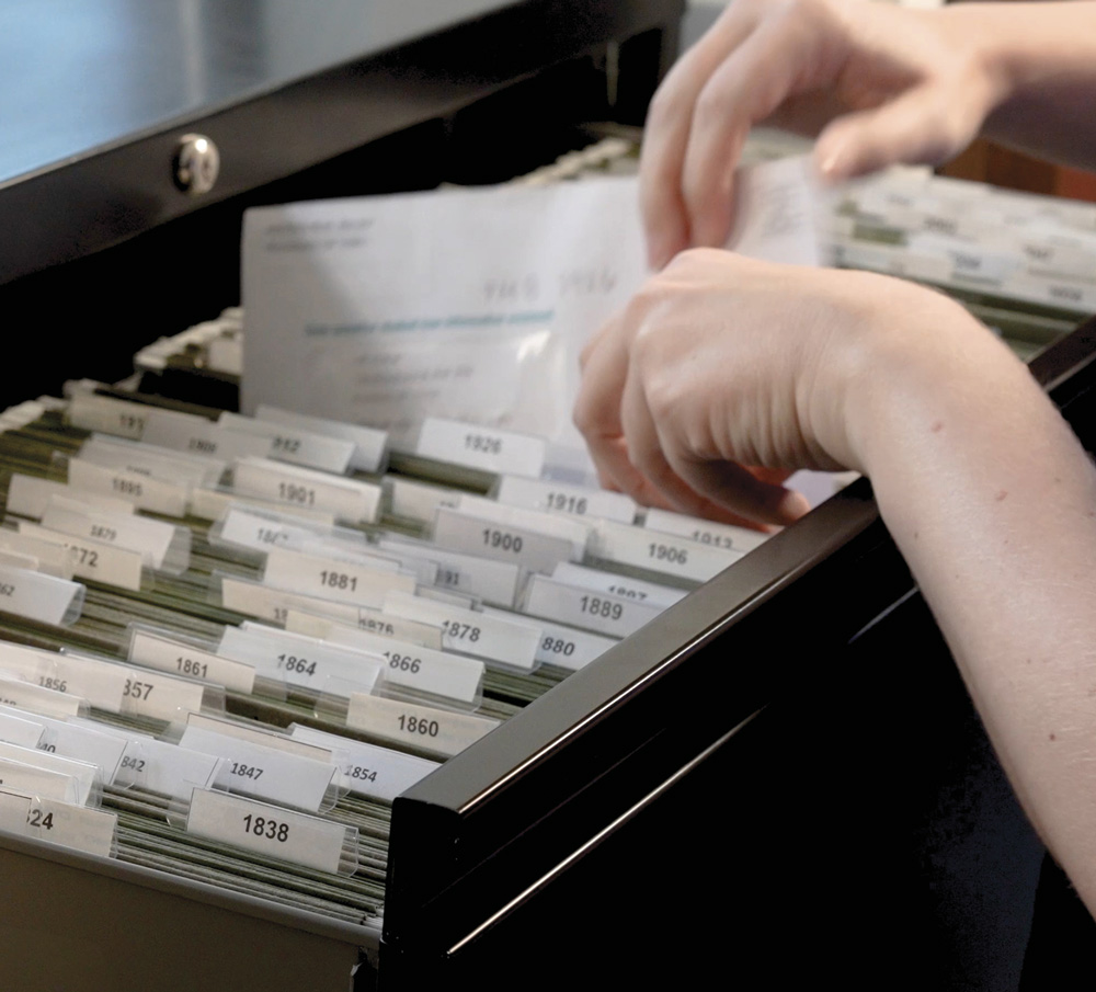 Hands organizing index cards in a filing cabinet with labels of years from the 1800s to 1900s.