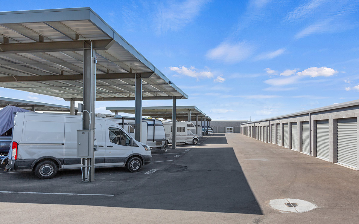 Outdoor facility with covered parking for vehicles and a long row of traditional drive-up storage units under a bright sky.