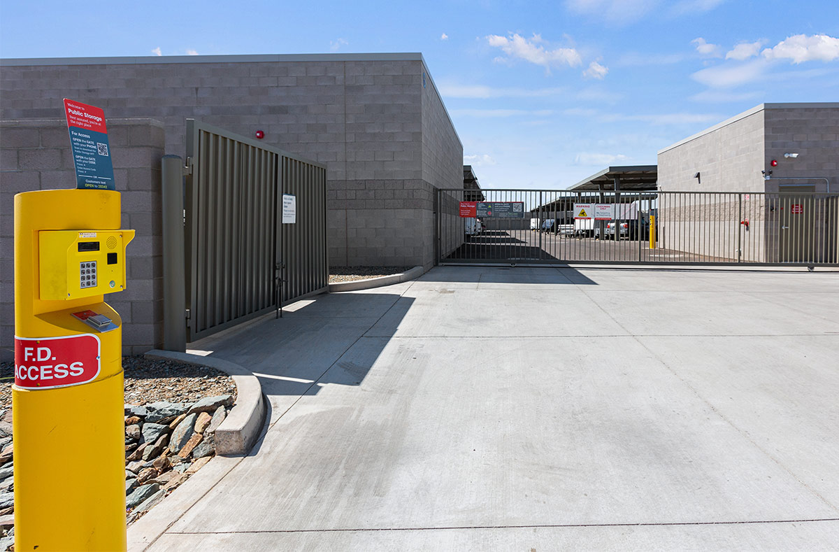 Main entrance gate of a storage facility between block buildings, with a yellow keypad and safety bollard for access.