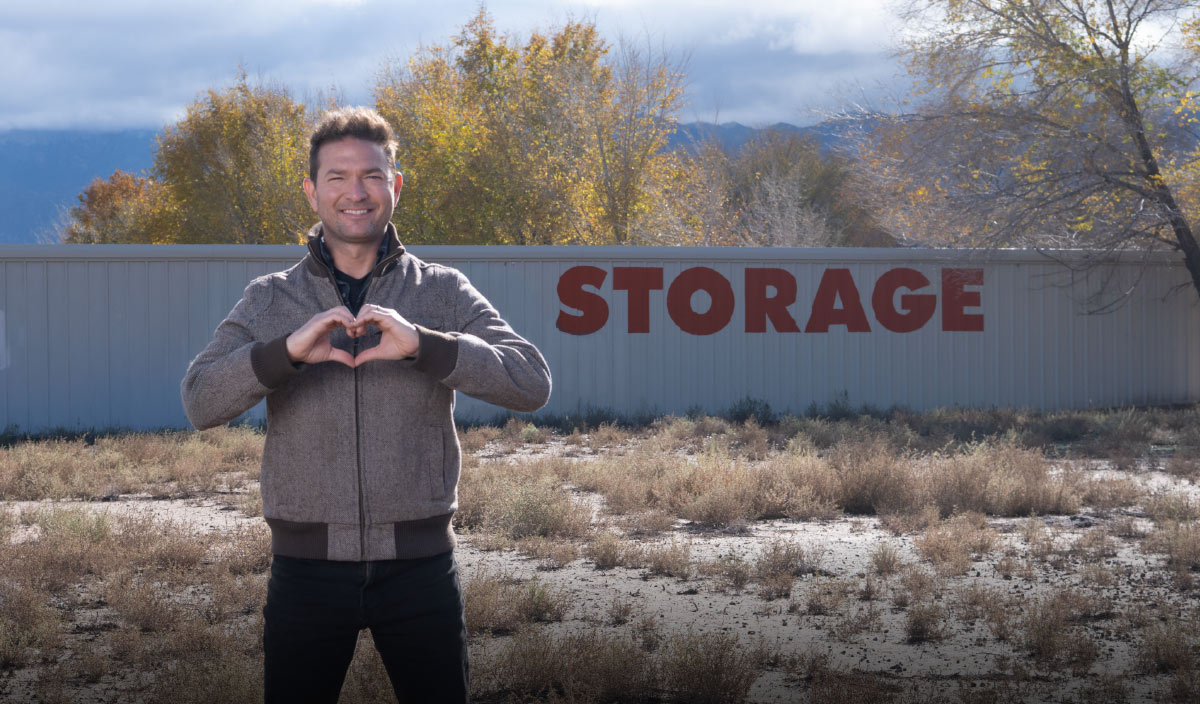 Man standing in a field making a heart shape with his hands in front of a long building with "STORAGE" painted in red.
