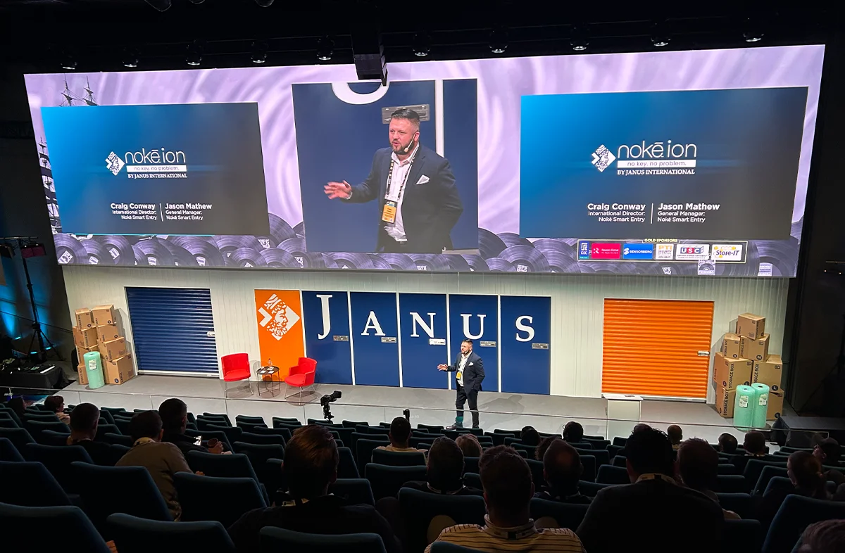 Keynote speaker presenting on a large stage featuring 'Noke Ion' and 'Janus' branding, with an audience seated in a theater-style auditorium.
