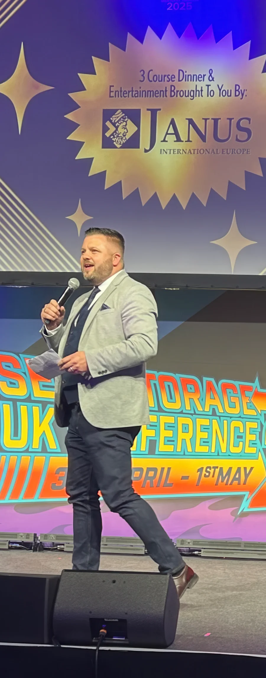 Keynote speaker in a grey blazer addressing the audience at a Janus International Europe sponsored 3-course dinner and entertainment event.