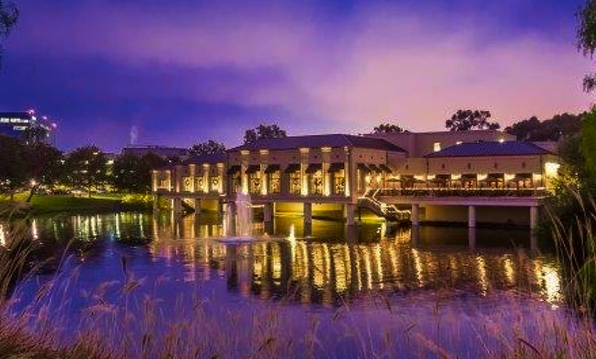 Exterior view of a luxury event venue at night with warm yellow lighting reflecting off a tranquil pond with a lit fountain.