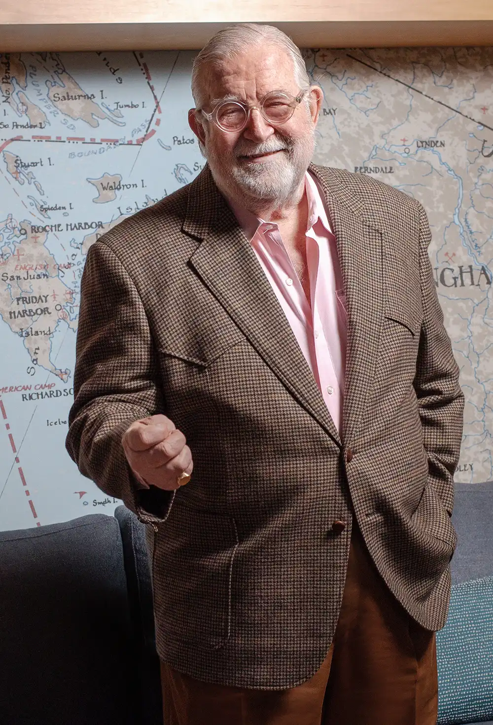 Portrait of Hardy Good, smiling while wearing round glasses, a pink shirt, and a brown checkered blazer. He stands in front of a large nautical wall map.