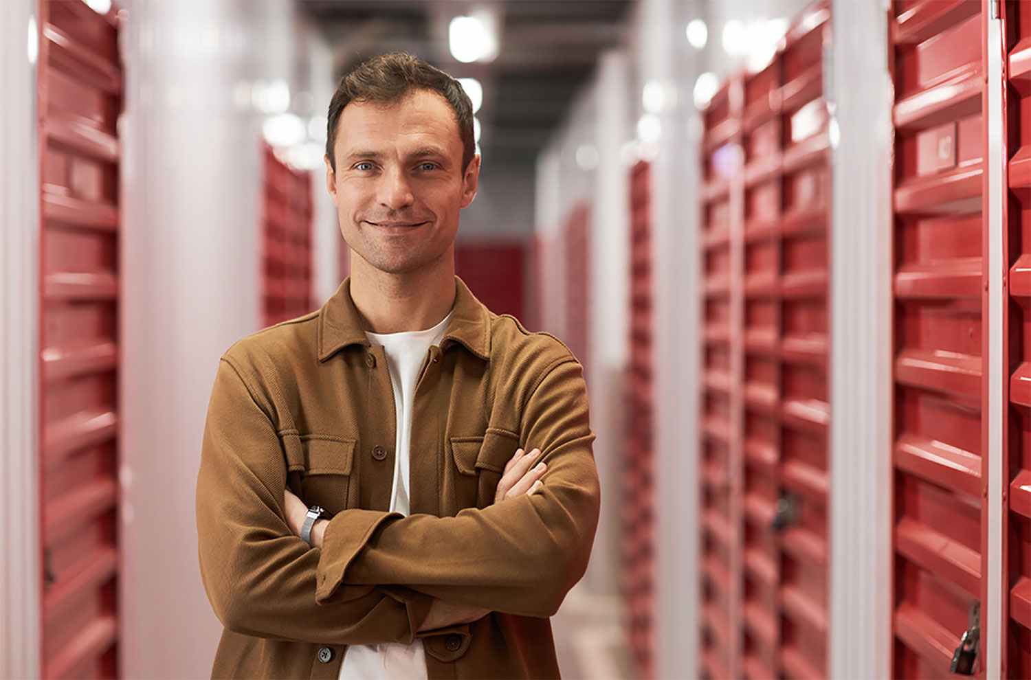 A smiling man with arms crossed standing in a bright indoor storage facility hallway lined with red unit doors.