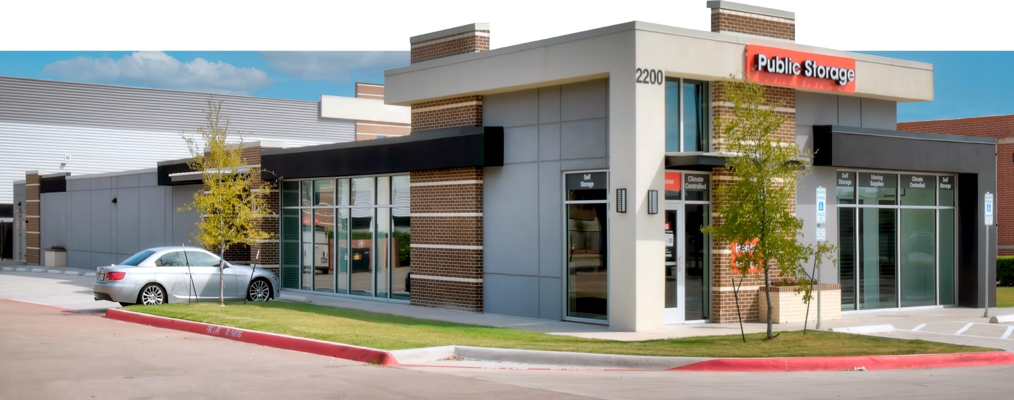 Wide exterior view of a modern Public Storage facility with a silver sedan parked in front of large glass windows and brick accents.