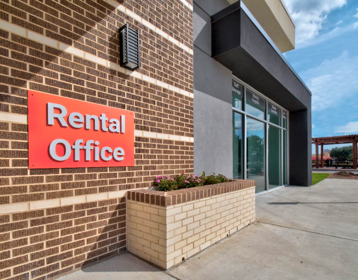A "Rental Office" sign mounted on a brick wall next to a modern glass-entry door.