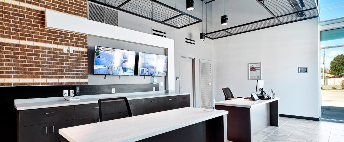 Alternative interior view of the rental office workspace showing desks, cabinetry, and a brick feature wall with security monitors.