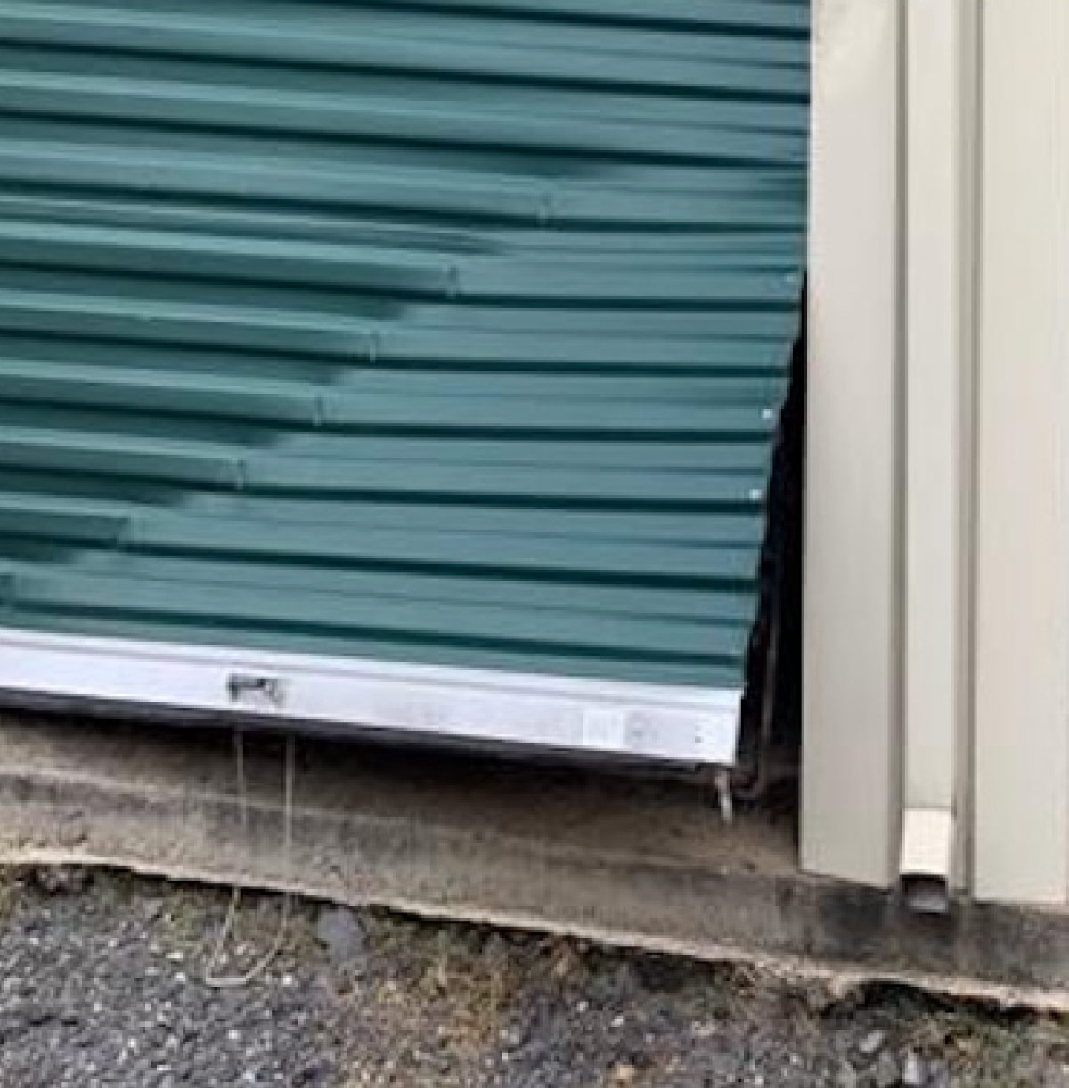 A damaged green corrugated metal roll-up door at a self-storage facility, showing significant denting and misalignment along the bottom track