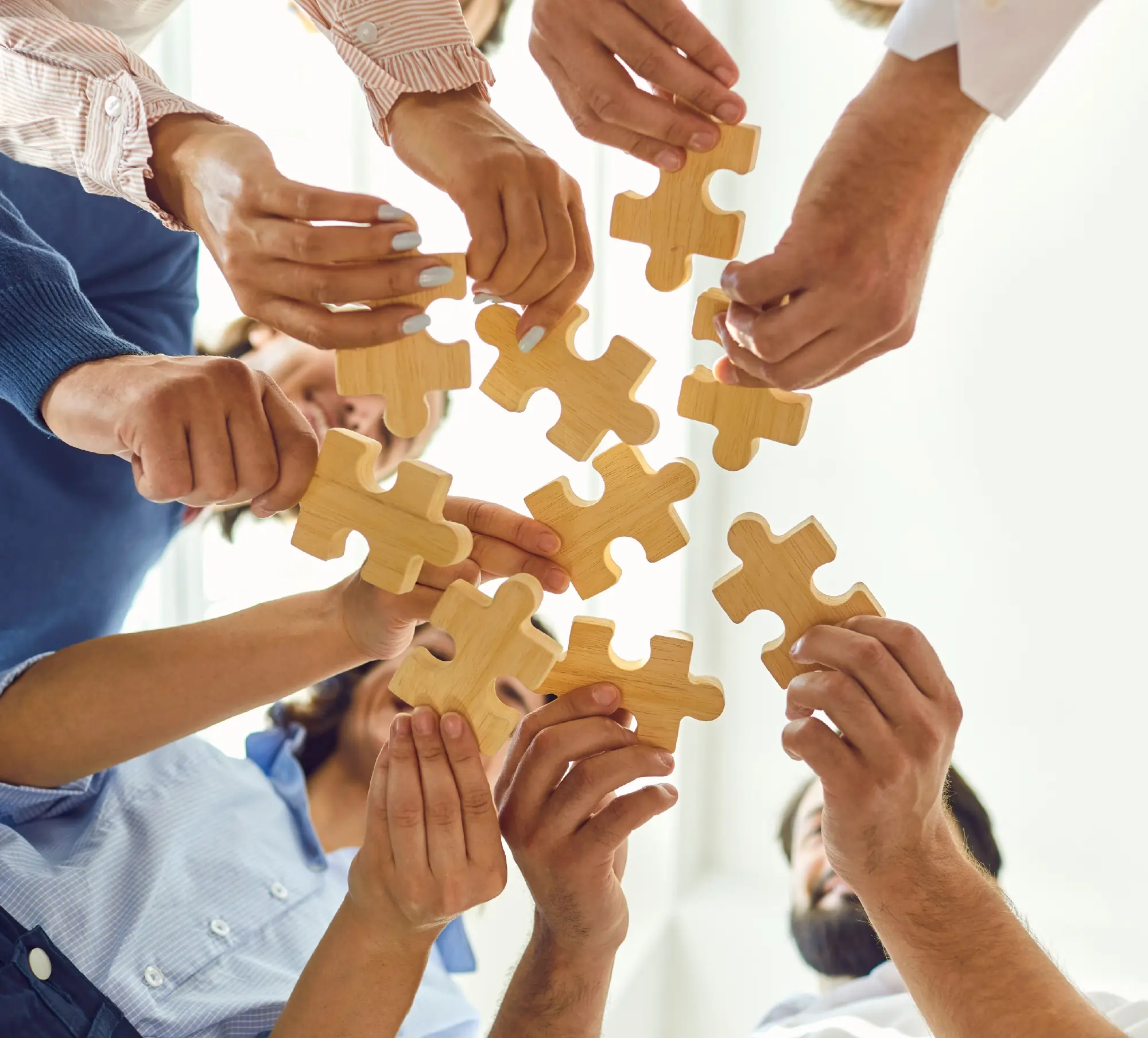 A low-angle shot looking up at a group of people standing in a circle, collectively holding and fitting together large wooden puzzle pieces against a bright background to symbolize teamwork and collaboration