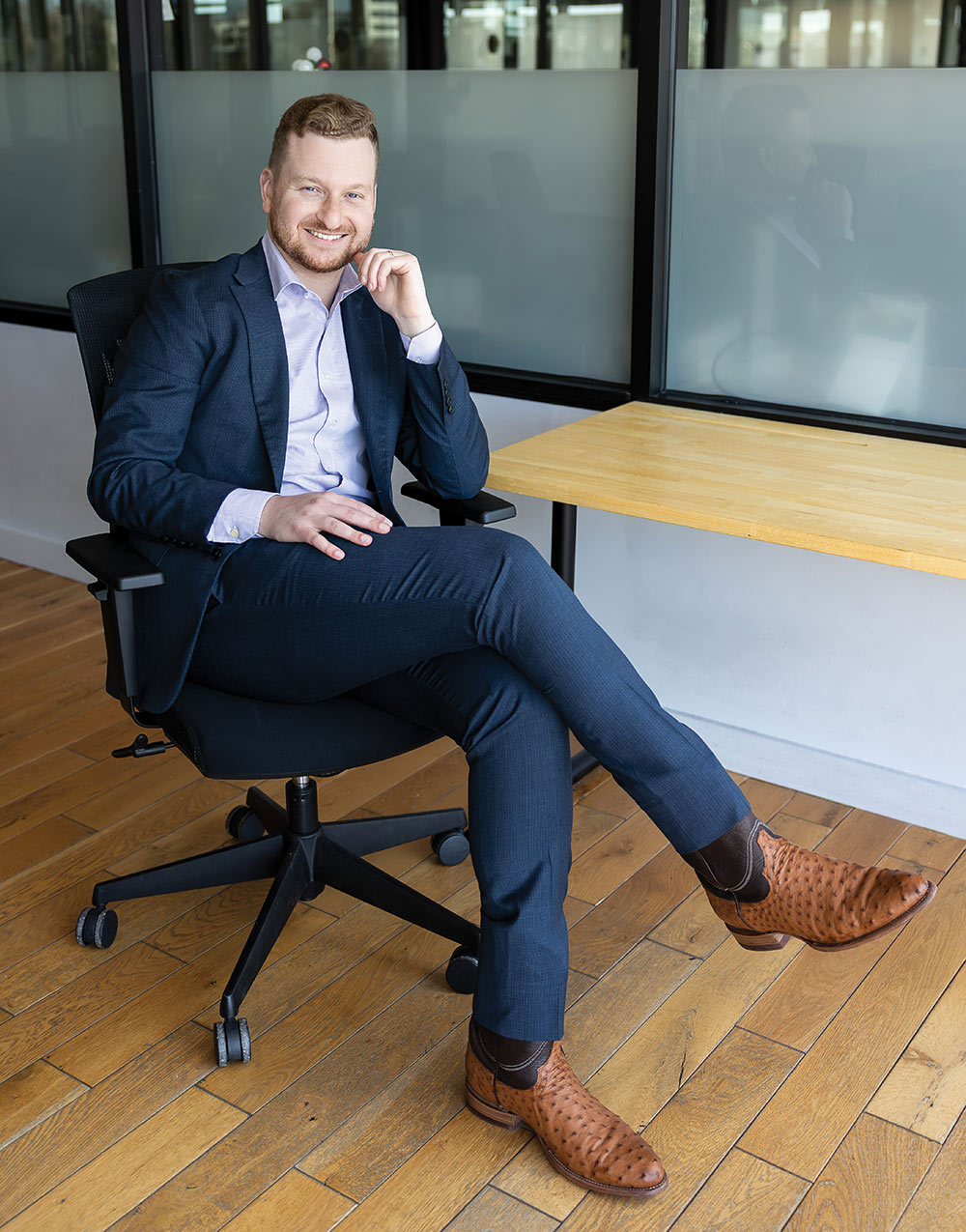 Noah Starr sitting on a desk chair wearing a navy suit and brown cowboy boots