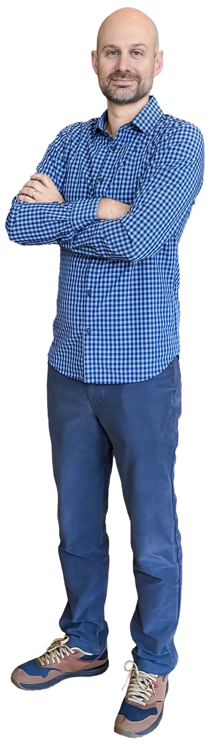 Full-length body portrait of Mitch Briggs, a smiling, bald man with a beard, standing with his arms crossed over his blue checkered button-down shirt, wearing blue trousers and brown sneakers