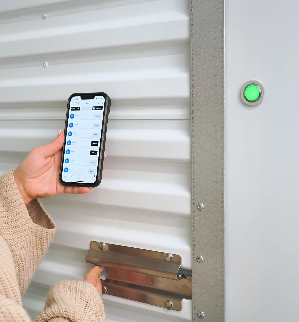 A customer using a smartphone app to wirelessly unlock a white corrugated metal storage unit door equipped with a Janus International electronic smart lock and a glowing green status indicator