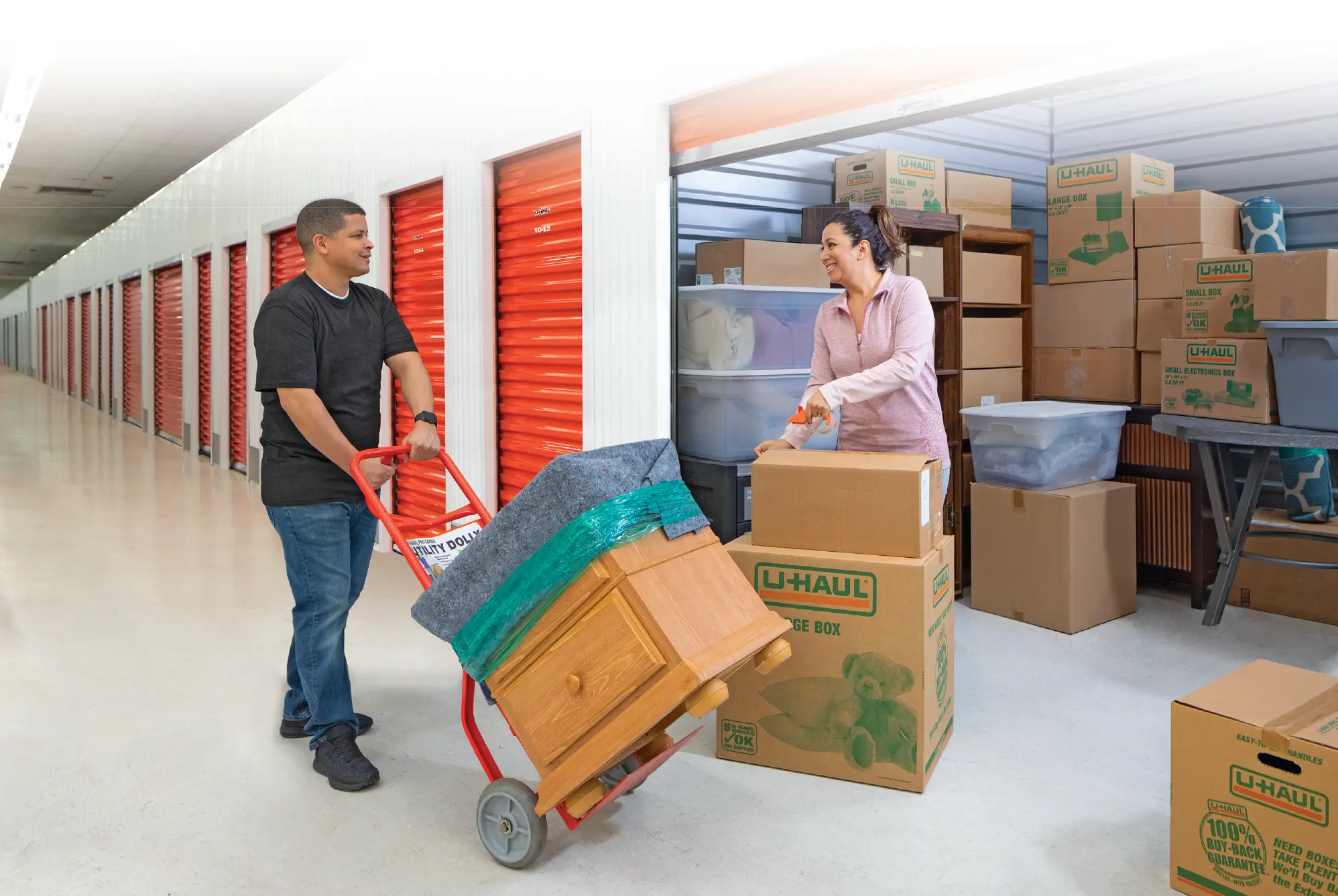 A man using a red utility dolly to move a wooden dresser into a clean, well-lit indoor U-Haul self-storage facility, while a woman organizes branded moving boxes inside a storage unit.