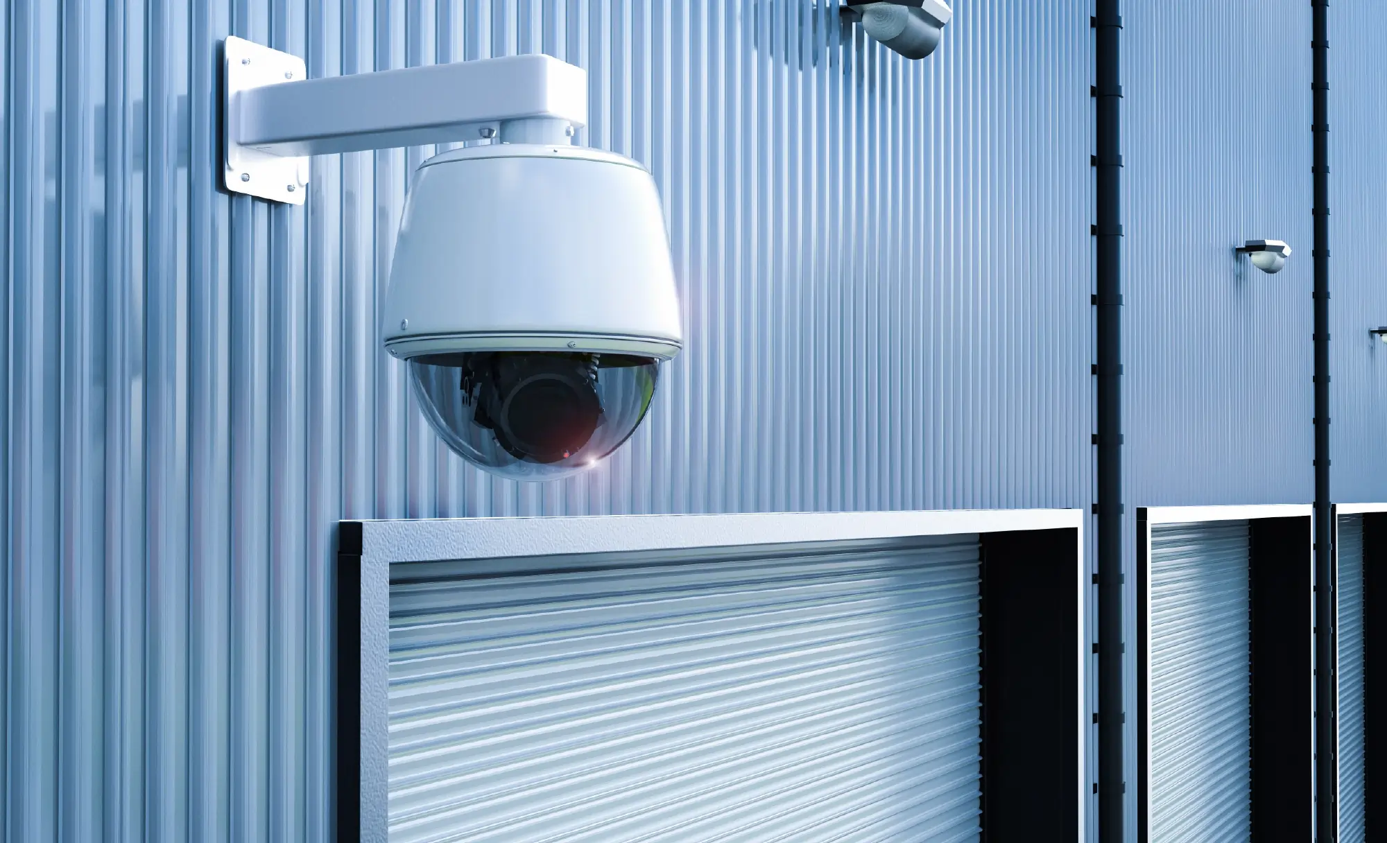 A high-angle shot of a white dome security camera mounted on the corrugated metal wall of a storage facility, positioned above a row of blue roll-up garage doors