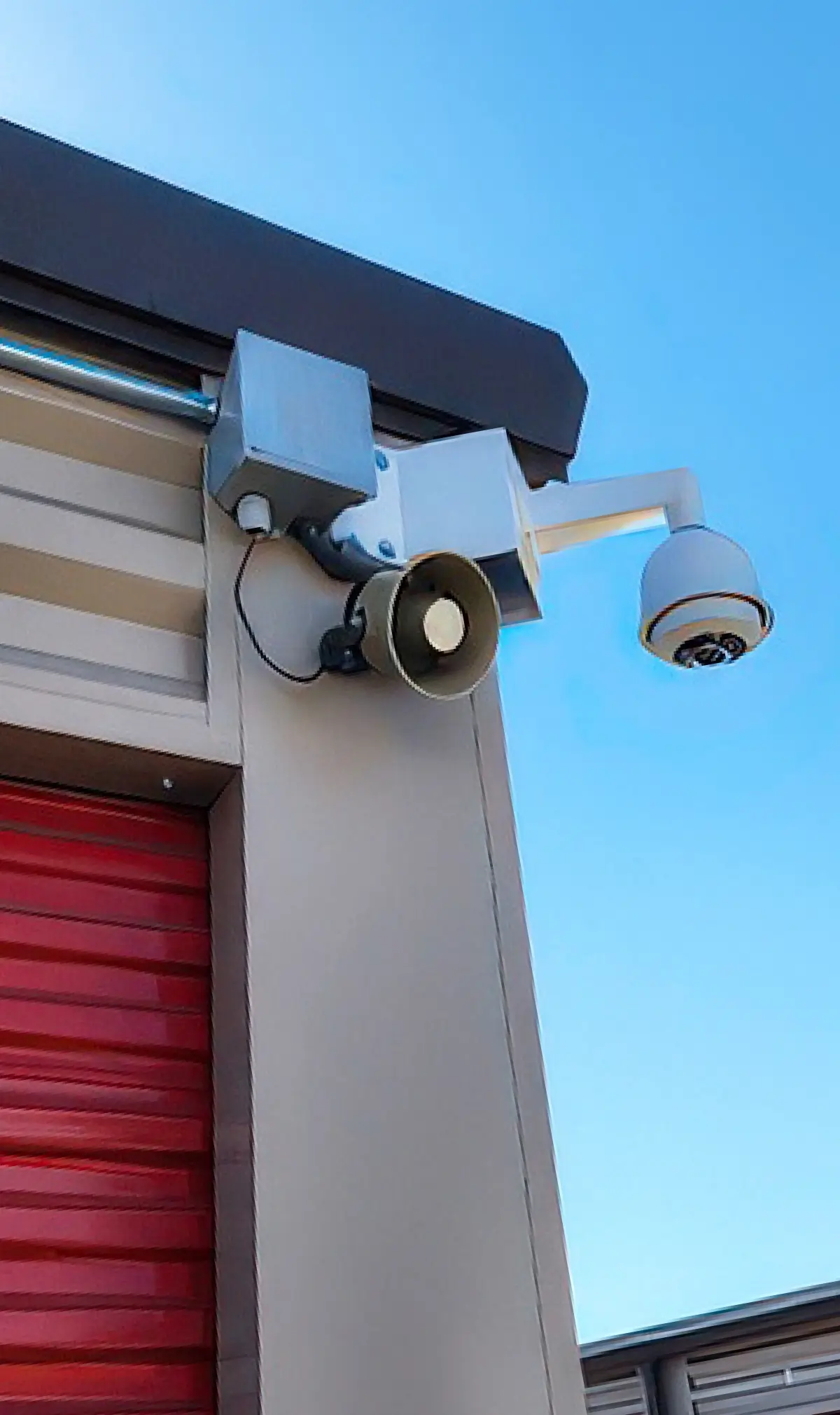 A low-angle shot of a white dome security camera and an attached loudspeaker mounted to the corner of a tan storage building; A red roll-up door is visible on the left under a clear blue sky