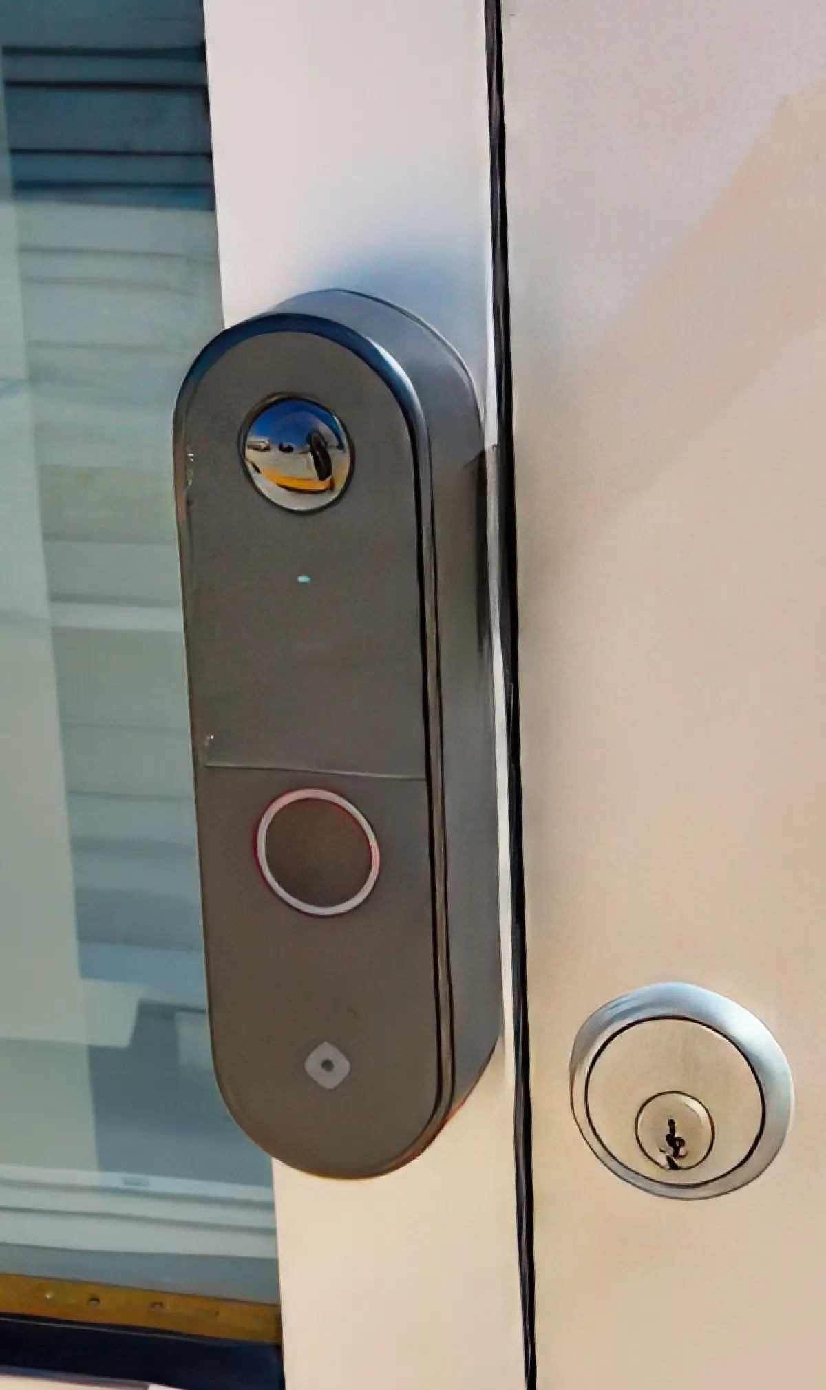 A close-up of a modern commercial glass door featuring a traditional key cylinder lock and a sleek dark grey smart video doorbell camera and or intercom system with a circular camera lens and a touch sensor