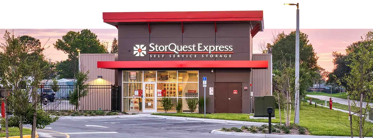 A wide landscape photograph of a StorQuest Express Self Service Storage facility at dusk; The building features a modern brown and tan exterior with bright red awnings, large glass windows showcasing an interior office, and a paved parking lot with green landscaping in the foreground