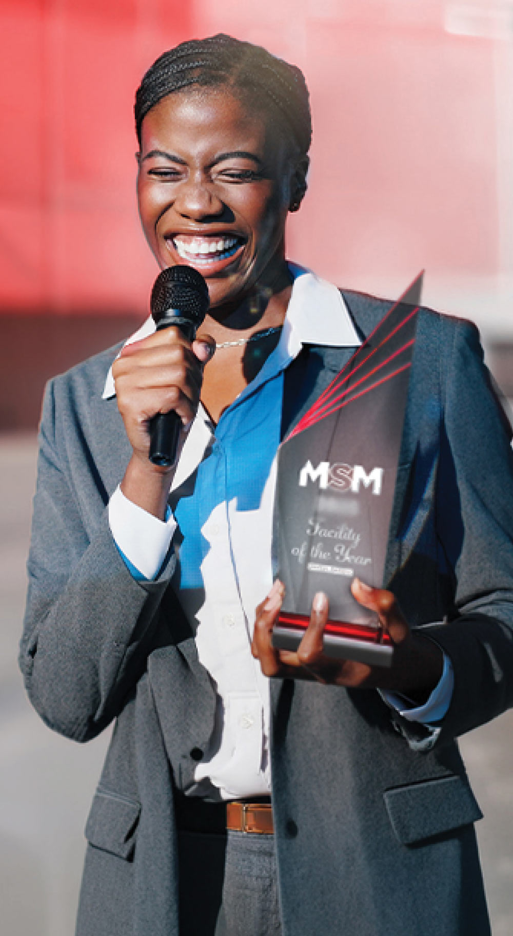 Person in a suit holding a microphone and a trophy, smiling outdoors.