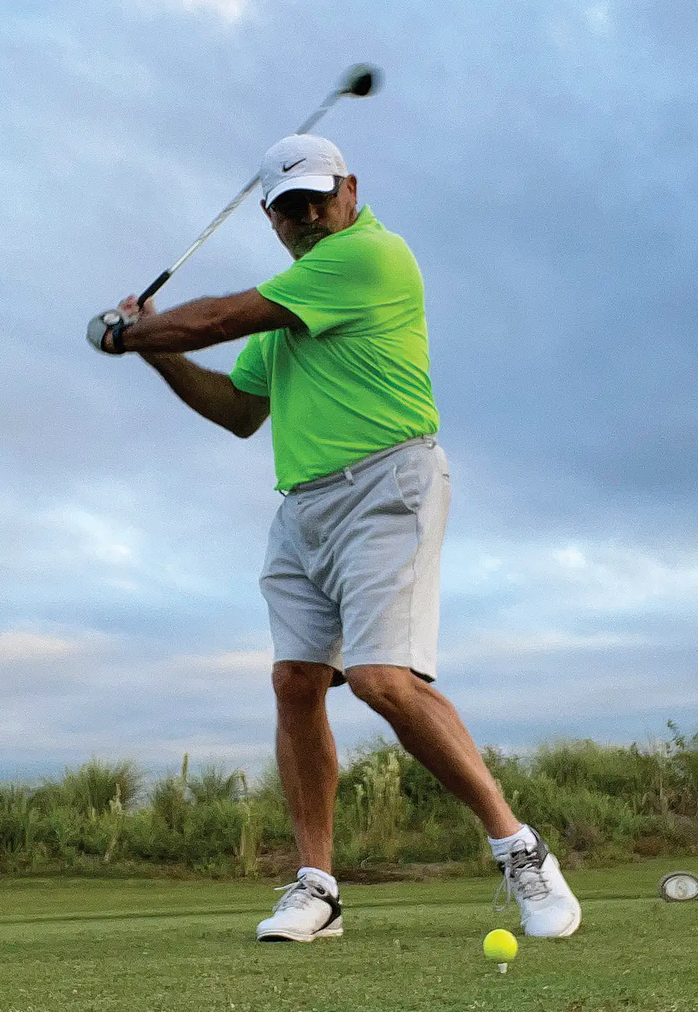 A man in a bright lime green shirt, white shorts, and a white baseball cap swings a golf club on a green course under a cloudy sky. A yellow golf ball sits on a tee in the foreground.